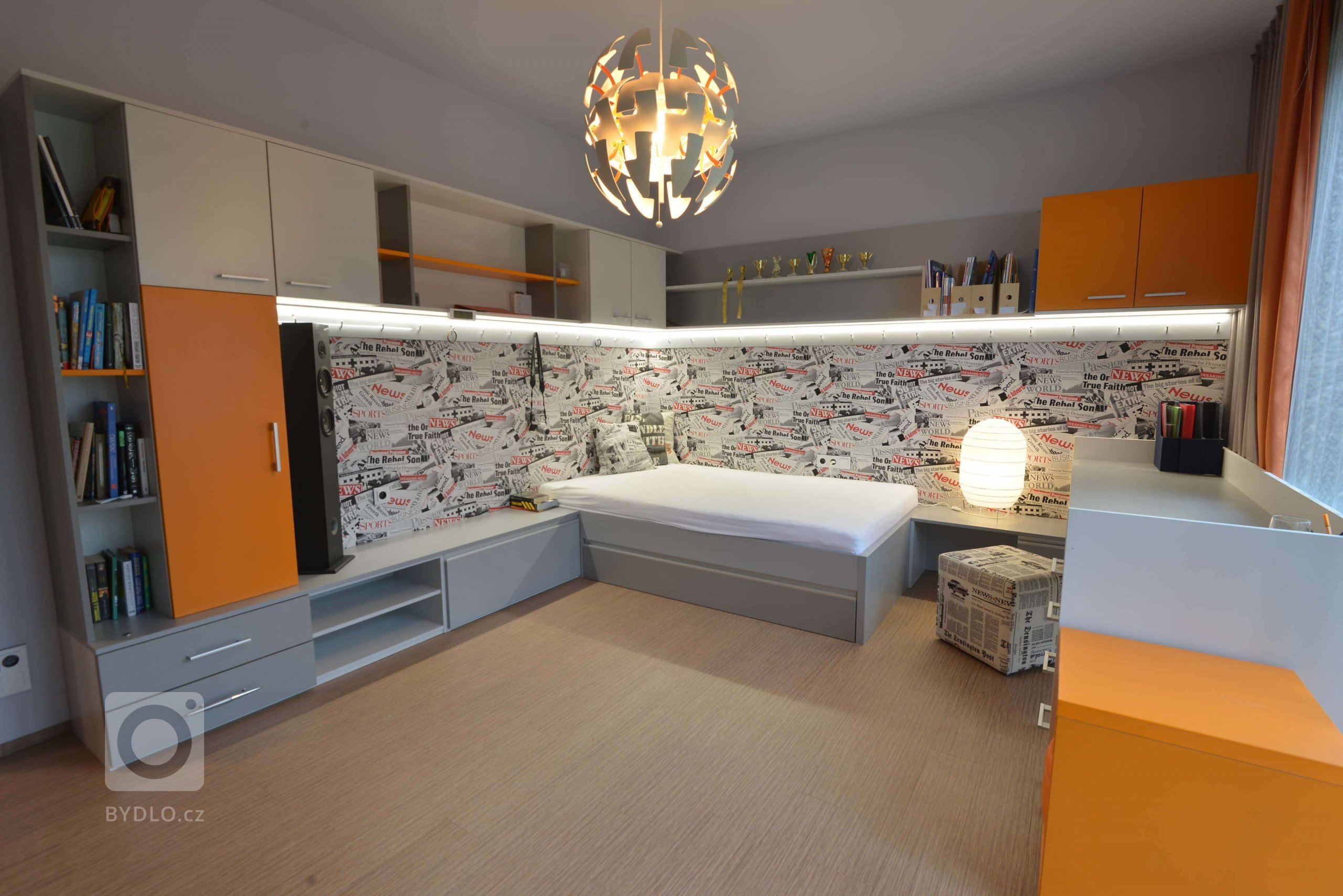Studentský pokoj pro chlapce. Barvy jsou po dohodě stříbrná v kombinaci s oranžovou. Okolo postele máme novinovou tapetu. Ve vrchních skříňkách je zabudované osvětlení LED se stmíváním. 
