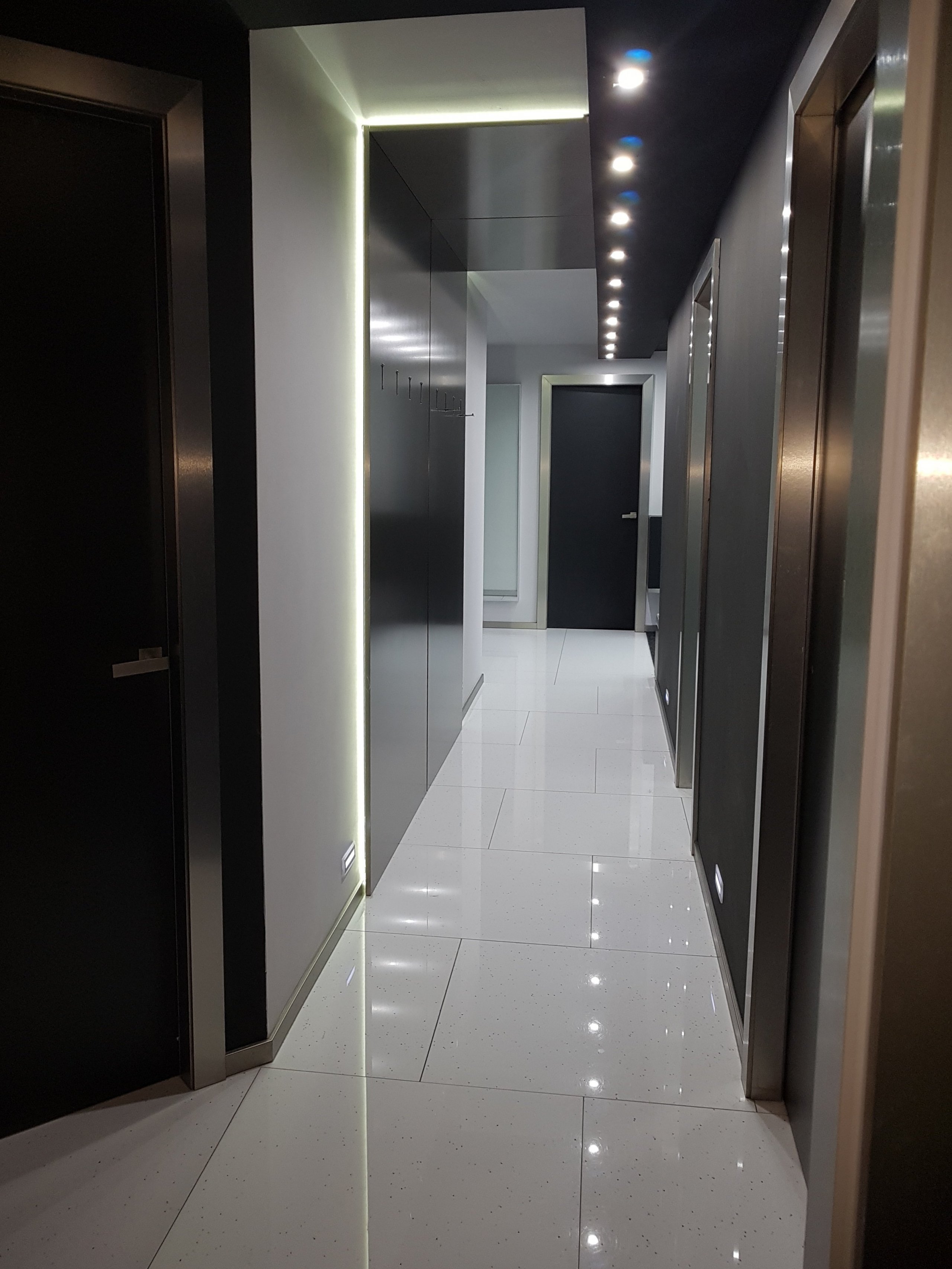 Dlouhá chodba nebyla moc široká, přes-to jsme do ní potřebovali umístit věšákovou stěnu. Je to věšení pro návštěvy, rodina by měla uklízet, taková byla úvaha.  Udělali jsme jí ze stříbrného kovolaminátu se sklopnými věšáčky. V případě že na ní nic nevisí je to velice čistý prvek se skrytým osvětlením, které přes strop přechází na druhou stranu a tvoří efektní dekoraci.  Protilehlá stěna je v černé barvě.  Dekorace na tuto stěnu, jsou stále ještě mezi uvažovanými  dodělávkami. 