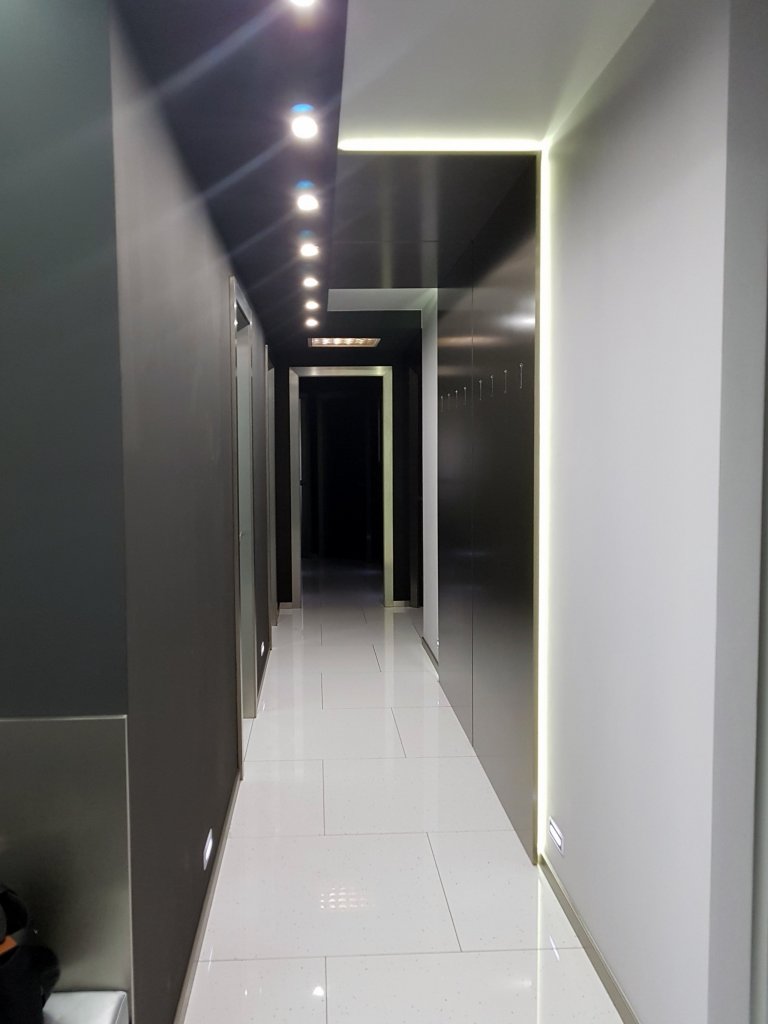 Pohled z druhé strany. Dlouhá chodba nebyla moc široká, přes-to jsme do ní potřebovali umístit věšákovou stěnu. Je to věšení pro návštěvy, rodina by měla uklízet, taková byla úvaha.  Udělali jsme jí ze stříbrného kovolaminátu se sklopnými věšáčky. V případě že na ní nic nevisí je to velice čistý prvek se skrytým osvětlením, které přes strop přechází na druhou stranu a tvoří efektní dekoraci.  Protilehlá stěna je v černé barvě.  Dekorace na tuto stěnu, jsou stále ještě mezi uvažovanými  dodělávkami.  