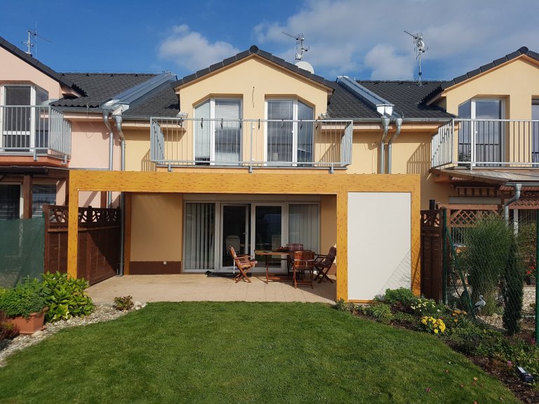 Lamelová pergola v imitaci dřeva a screenové rolety v husté zástavbě.
