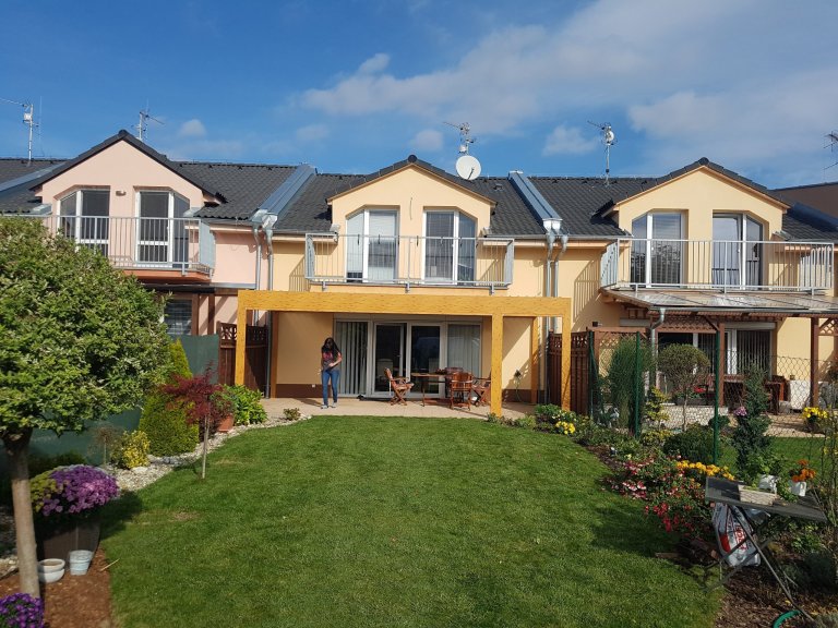 Lamelová pergola v imitaci dřeva a screenové rolety v husté zástavbě.
