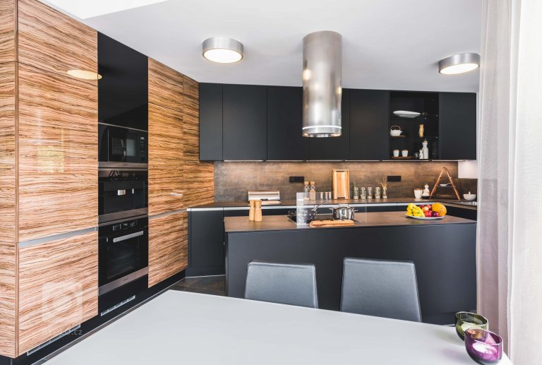 Luxusní tmavá dvířka Top Akryl Matt Black navazují na skříně s vysokým leskem v dřevěném dezénu Oliva, díky čemuž místnost získává neutuchající energii a…