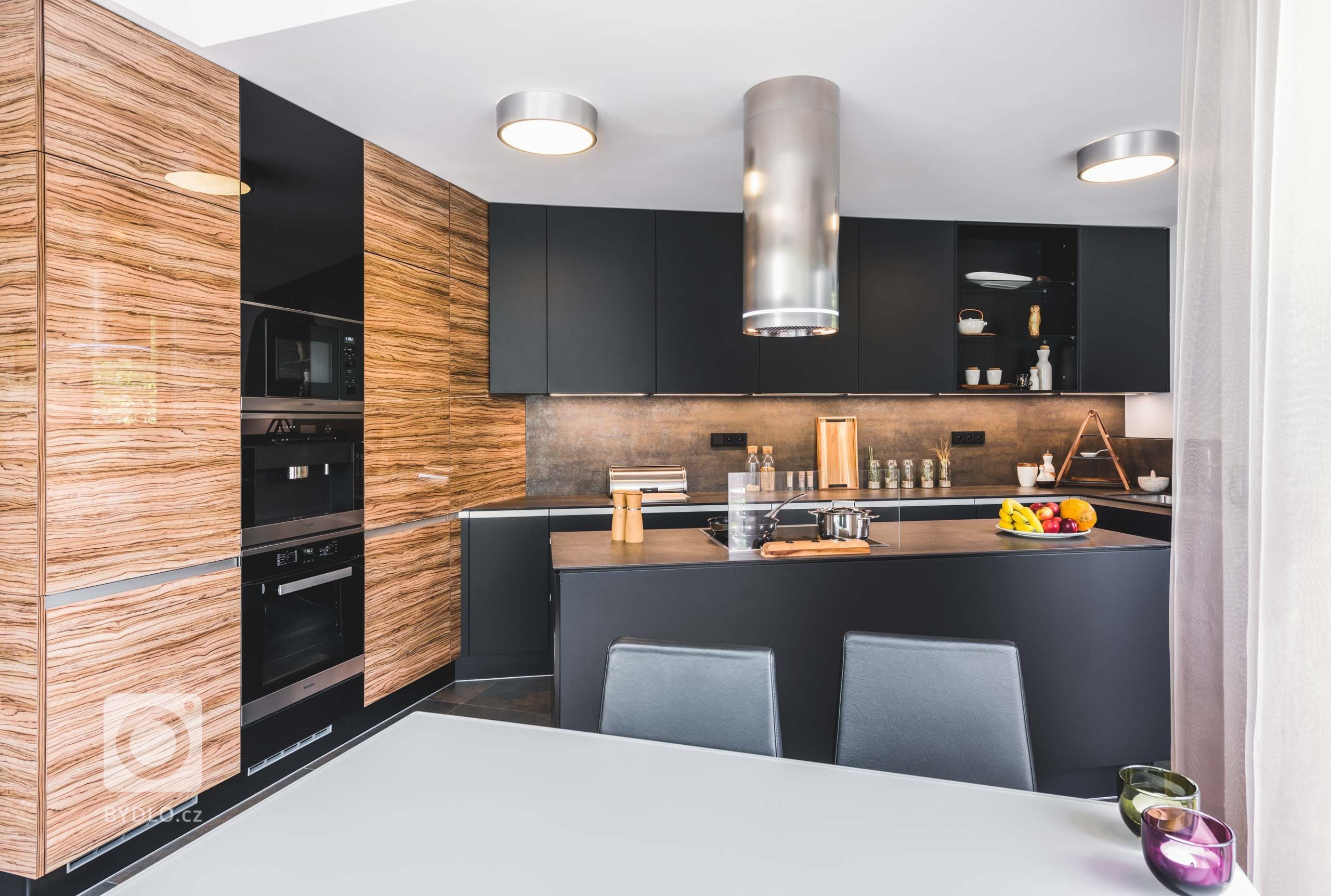 Luxusní tmavá dvířka Top Akryl Matt Black navazují na skříně s vysokým leskem v dřevěném dezénu Oliva, díky čemuž místnost získává neutuchající energii a…