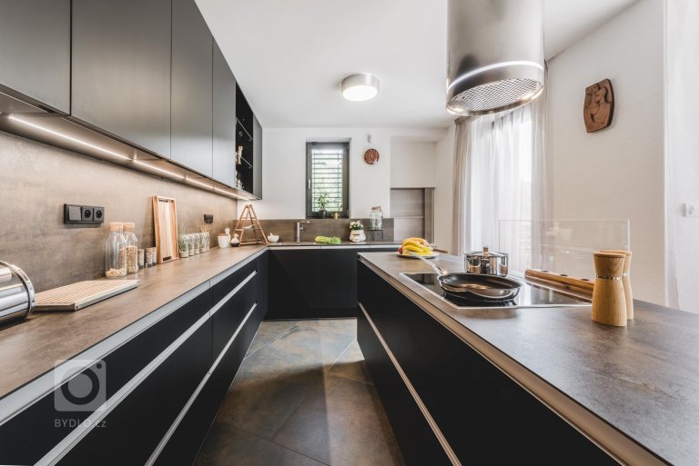 Luxusní tmavá dvířka Top Akryl Matt Black navazují na skříně s vysokým leskem v dřevěném dezénu Oliva, díky čemuž místnost získává neutuchající energii a…