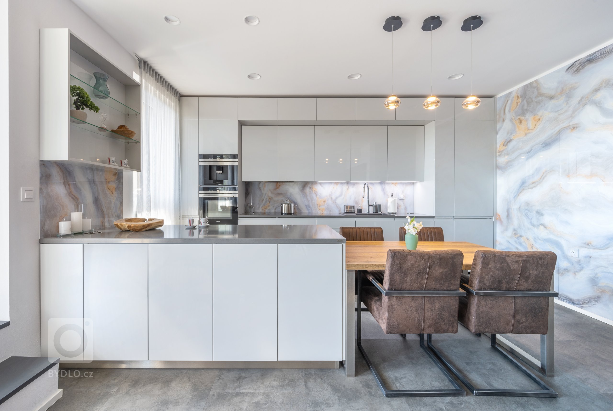 Sestava samotná využívá symetrii horních a dolních skříněk v provedení Light Grey. Bílé, leskle lakované plochy dvířek jsou okouzlující na pohled i vhodné na každodenní provoz. Ve spojení se zástěnami působí vzdušně a přidávají prostoru na velikosti. Zafrézované úchytky Pure už tak nepřehlédnutelnou hladkost a čistotu linií ještě vyzdvihují.